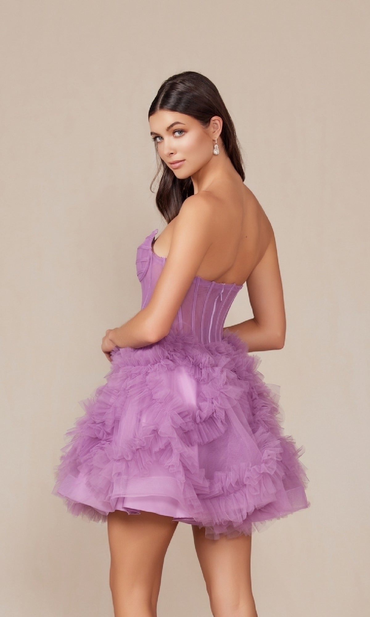 A woman in the Short Strapless Ruffled Homecoming Dress Y859, a lavender mini, stands partially turned away from the camera and looks over her shoulder against a plain beige background.
